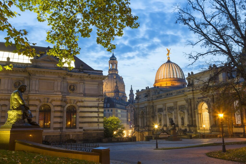 Illuminated town square with facades of domed church, college and sculptures.