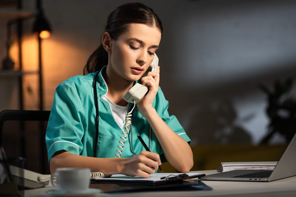 attractive nurse in uniform talking on telephone and writing in clipboard during night shift