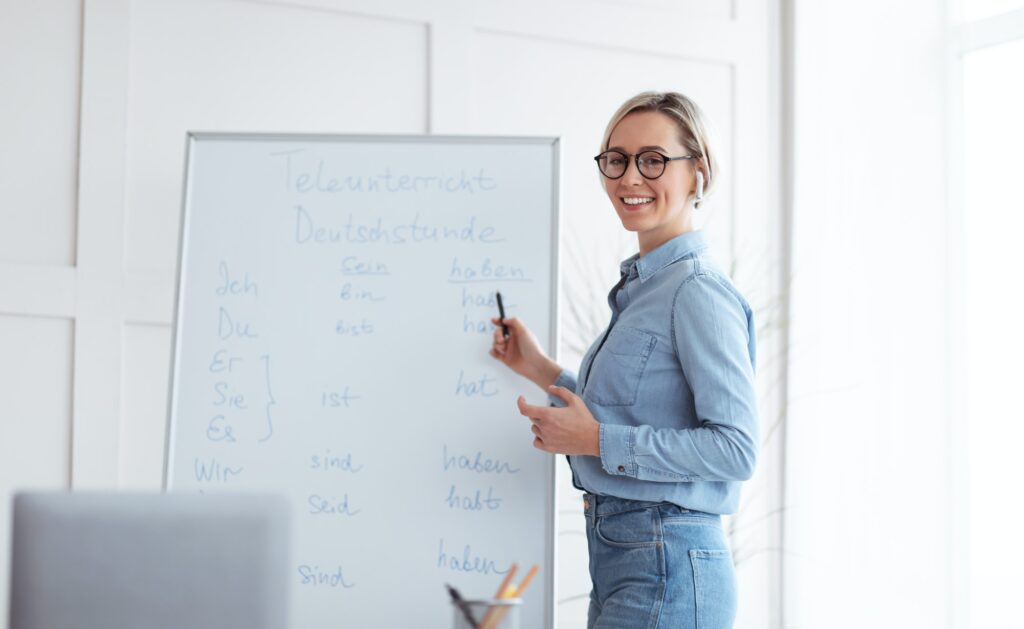 Happy young teacher standing near blackboard and explaining German lesson to students online from